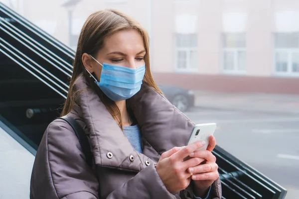 Cerrahi maske takan beyaz bir kadın şehrin dışında yürürken akıllı telefon kullanıyor.