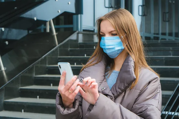 Cerrahi maskeli genç kadın şehirde yürürken akıllı telefon kullanıyor.