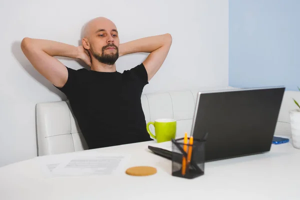 Thouthful uomo calvo con barba in t-shirt nera che lavora sul suo taccuino a casa, vista laterale — Foto Stock