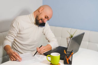 Beyaz balıkçı yakalı, sakallı, gülümseyen kel adam defter üzerinde çalışıyor ve telefonda konuşuyor.