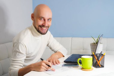 Beyaz balıkçı yakalı, sakallı, gülümseyen kel adam defter üzerinde çalışıyor.