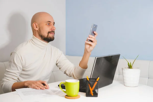 Beyaz balıkçı yakalı, sakallı, gülümseyen kel adam evde defter üzerinde çalışıyor ve telefon, yan görüş arıyor.