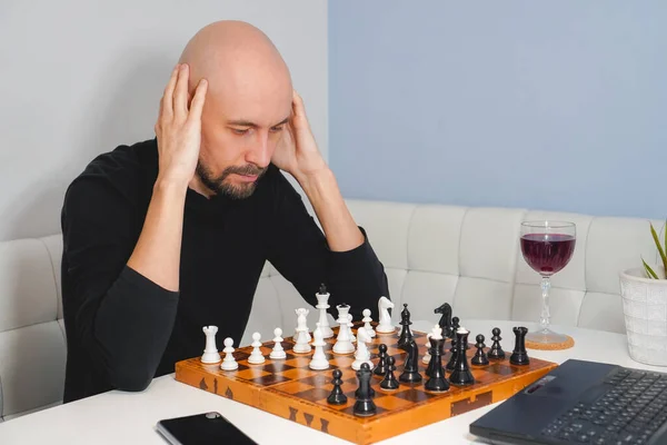 Figyelmes, kopasz, szakállas férfi sakkozik a fedélzeten laptop és borosüveg közelében. Stratégia és versenykoncepció. Oldalnézet — Stock Fotó