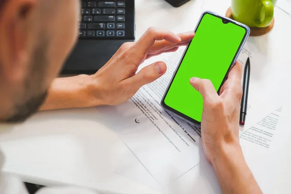 Mockup image, muž ruka drží prázdné zelené obrazovce mobilní chytrý telefon, pracuje na notebooku Royalty Free Stock Obrázky