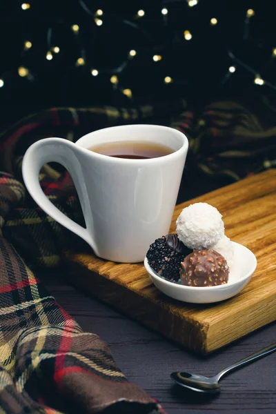 Taza blanca de té con cuchara y caramelos de chocolate sobre fondo borroso con luces de Navidad — Foto de Stock