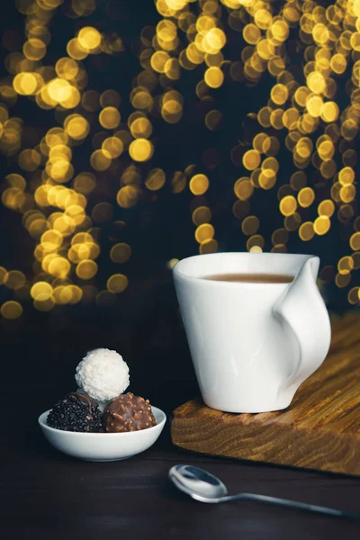 Taza blanca de té con cuchara y caramelos de chocolate sobre fondo borroso con luces de Navidad — Foto de Stock