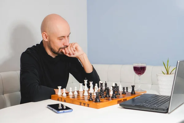 Задумчивый лысый мужчина с бородой играет в шахматы на борту рядом с ноутбуком и бокалом вина. Концепция стратегии и конкуренции. Вид сбоку — стоковое фото