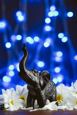 Mavi arka planda papatya çiçekli bir fil figürü. Papatyalar arasındaki siyah taş fil heykelciği