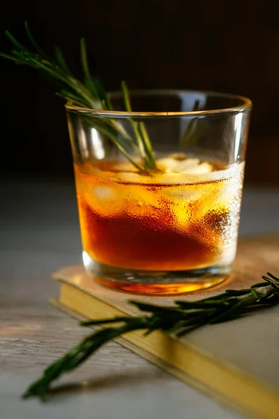 Whisky kyselý koktejl s rumovým ledem a rozmarýnem v broušené sklenici na knihu. Alkoholový koktejl v paprscích světla Stock Snímky