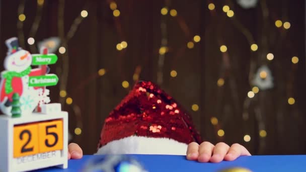 Petite fille souriante au chapeau de Père Noël regardant par derrière la table. Enfant à Noël — Video