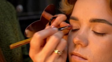 Profesyonel makyöz, genç bir kadının göz kapağına göz farı sürüyor. Kapat.