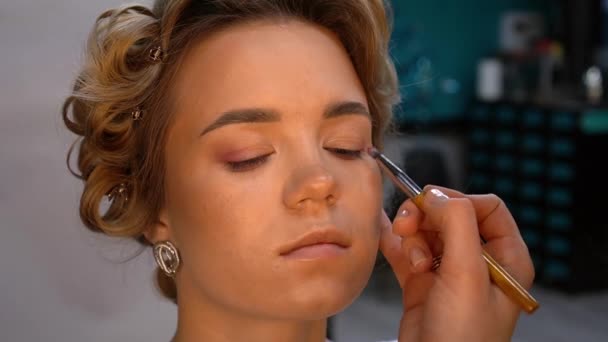 O artista de maquilagem profissional aplica a sombra de olho à pálpebra da mulher jovem. Fechar — Vídeo de Stock
