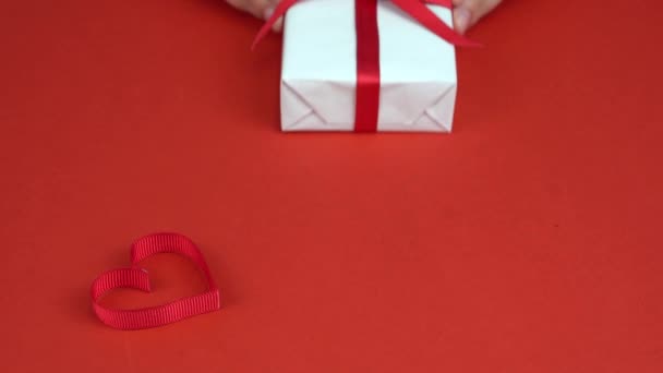 Le mani di ragazza mettono la scatola regalo bianca con fiocco rosso su tavolo rosso decorato con cuore. San Valentino — Video Stock