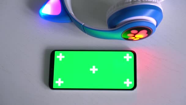 Smartphone con pantalla verde en blanco maqueta con auriculares inalámbricos para niños aislados sobre fondo gris — Vídeos de Stock