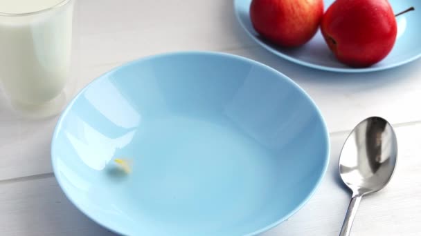 Кукурузные хлопья выливают в голубую чашу на деревянный стол. Кукурузные хлопья молоко и яблоко здоровый завтрак — стоковое видео