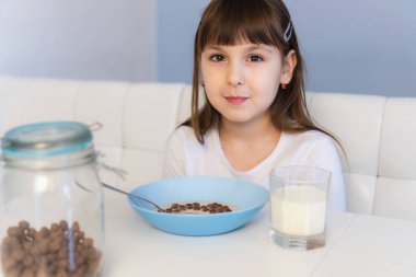 Küçük kız beyaz masada oturmuş sütlü çikolatalı mısır topu yiyor. Çocuk kahvaltı yapıyor.