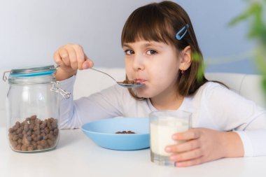 Küçük kız beyaz masada oturmuş sütlü çikolatalı mısır topu yiyor. Çocuk kahvaltı yapıyor.