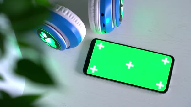 Gri arka planda izole edilmiş kablosuz kulaklıklı, boş yeşil ekran modeli olan akıllı telefon. — Stok video