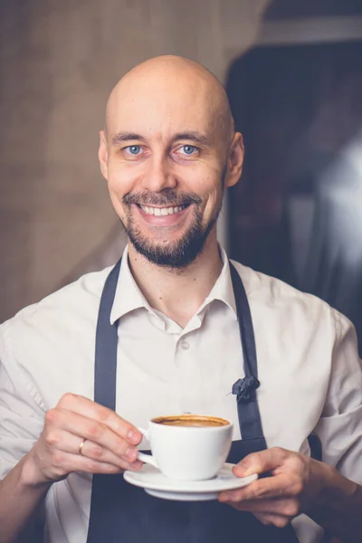 Usmívající se muž v zástěře drží šálek s kávovým kapučínem. Barista připravil čerstvou horkou kávu Stock Obrázky