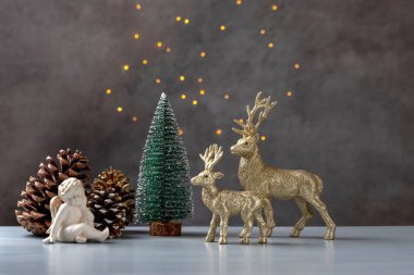Noel kompozisyonu. Melek, kozalak, ren geyiği ve beyaz bir masada Noel ağacı ve peri ışıklarıyla dolu gri bir arka plan..