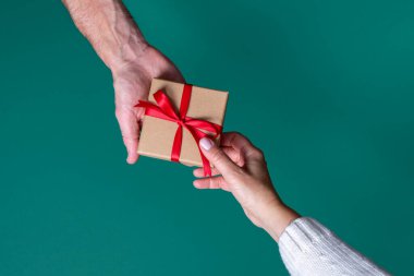 Erkek ve kadın ellerinin yeşil arka planda kırmızı kurdeleli hediye kutusunu tutuşu. Doğum günü hediyesi, Sevgililer Günü, Noel, Yeni Yıl. Boşluğu kopyala, üst görünüm.