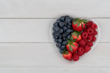 Sağlıklı karışık meyveler, çilek, ahududu, böğürtlen, kalp şeklinde bir kasede beyaz bir masada, üst manzara, kopyalama alanı.