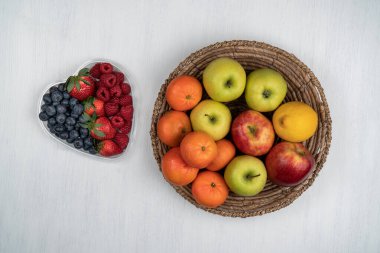 Sağlıklı karışık meyveler, çilek, ahududu, kalp şeklinde bir kasede yabanmersini ve beyaz bir masada elma ve mandalina dolu bir sepet..