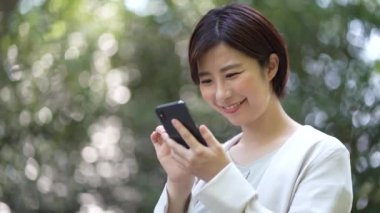 Zeki telefon kullanan ve parktaki kameraya gülümseyen neşeli Asyalı kadın.