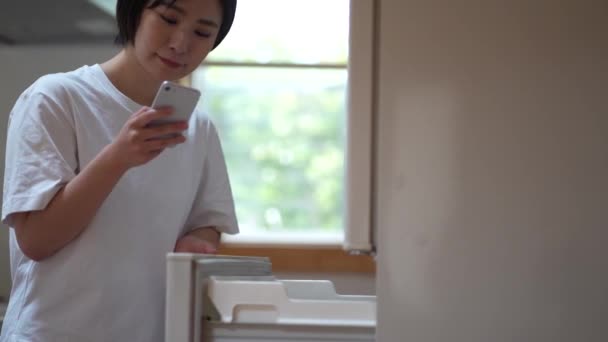 Uma Mulher Procurando Receitas Seu Smartphone Enquanto Olha Dentro Geladeira — Vídeo de Stock