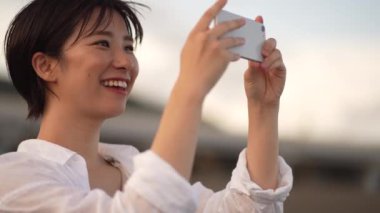 Gülümseyen Asyalı genç bir kadın gün batımında sahilde akıllı telefonuyla fotoğraf çekiyor.