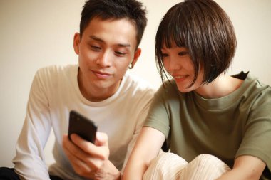 Asyalı çift evdeki koltukta otururken cep telefonu kullanıyor.