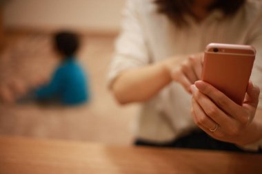 Genç Asyalı anne akıllı telefon kullanıyor, çocuk arka planda.