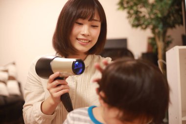 Asyalı anne oğlunun saçını kurutma makinesiyle kurutuyor. 