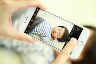 Erkek bebek yatakta yatıyor, anne akıllı telefonuyla fotoğraf çekiyor.