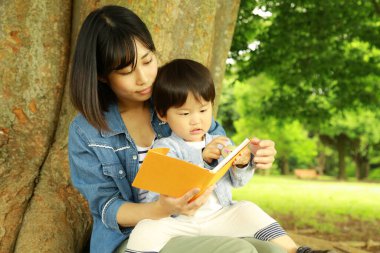 Genç Asyalı anne ve çocuğu parkta kitap okuyor.  