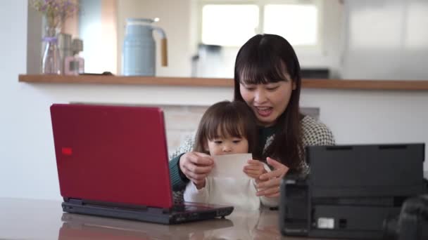 若いアジア系のお母さんと可愛い娘がノートパソコンとプリントカードを使って一緒に家で — ストック動画