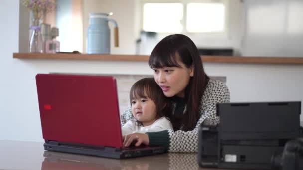 若いアジア系のお母さんと可愛い娘がノートパソコンとプリントカードを使って一緒に家で — ストック動画