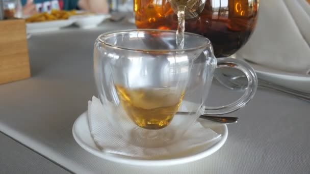 Transparante mok staat en vervolgens hete ceylon thee wordt gegoten in het van glazen theepot. Ober in het restaurant. — Stockvideo