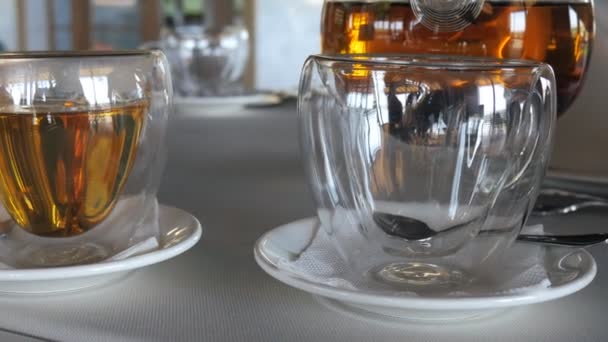 Transparent mug stands and then hot ceylon tea is poured into it from glass teapot. Waiter at the restaurant. Two cups of black tea. — Stock Video