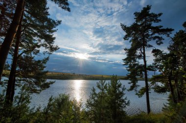 Gölün kıyısındaki çam ormanı ve bulutların arasından geçen suyun üzerindeki güneş.