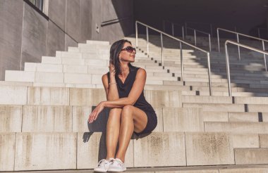 Siyah elbiseli, siyah saçlı kadın beton merdivenlerde poz veriyor. Kadın siyah güneş gözlüğü takıyor. Güzel, güneşli bir gün..