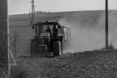 Traktör çayırın yanındaki yolda gidiyor. Traktör arkasında tozlu bir bulut oluşturuyor. Fotoğraf siyah beyaz..