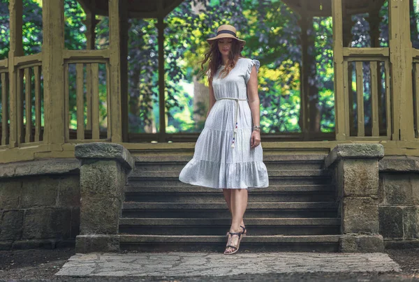 woman goes up the stairs that lead to the gazebo. The woman is wearing a white long dress, black sandals and has a hat on her head. There are trees behind the gazebo and nature is all around.
