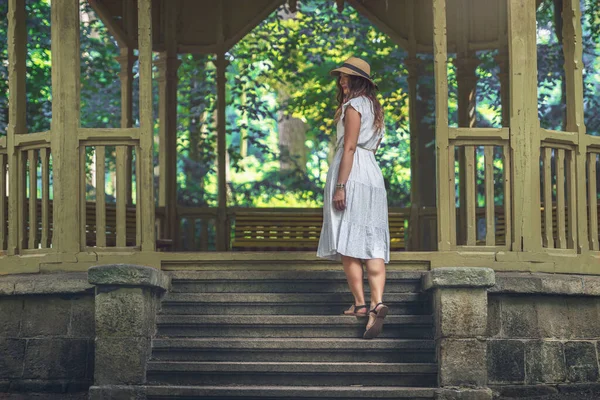 woman goes up the stairs that lead to the gazebo. The woman is wearing a white long dress, black sandals and has a hat on her head. There are trees behind the gazebo and nature is all around.