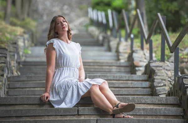 Woman sitting on the steps in a long white dress enjoying a summer day. The stairs lead through the park and are lined with railings. The woman has slippers on her feet.