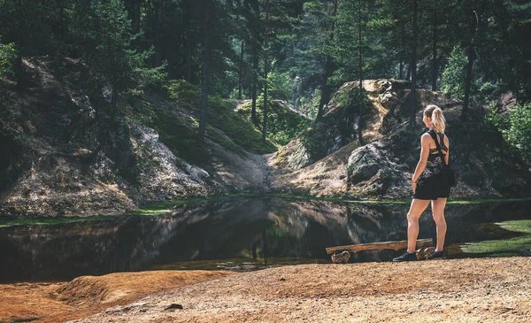 The girl is standing on the edge of the pond by the bench and looking towards the landscape beyond the pond. Behind the lake there is a forest and small rocks. There is red clay in front of the pond.