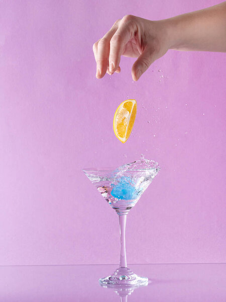 Woman hand throwing lemon into martini glass full with alcohol drink with splashes, against pastel pink background. Minimal party concept. Creative martini idea.