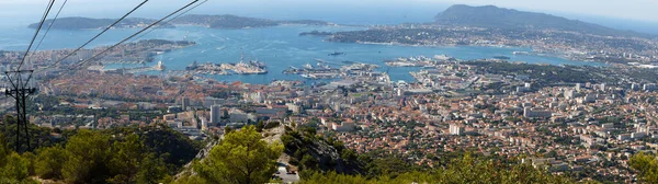 A scenic view of the city of Toulon from a hill called Mount Faron . Houses and buildings have red tiled roofs and big ships are parked in the harbor.