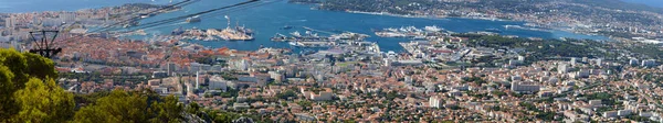 A scenic view of the city of Toulon from a hill called Mount Faron . Houses and buildings have red tiled roofs and big ships are parked in the harbor.