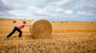 Genç bir kadın tarlalarda kuru saman balyalarını itiyor.